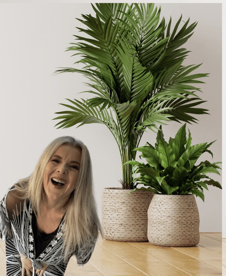 Image of Nadine Hill, founder of Glow Within and Yoga by Nadine Hill, a white woman with straight blonde hair, wearing a black and white kimono and laughing with green plants in the background.